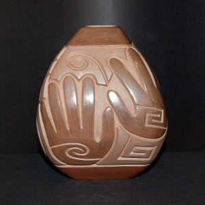 Spiral, hand and geometric design carved into a brown on brown jar with a square opening